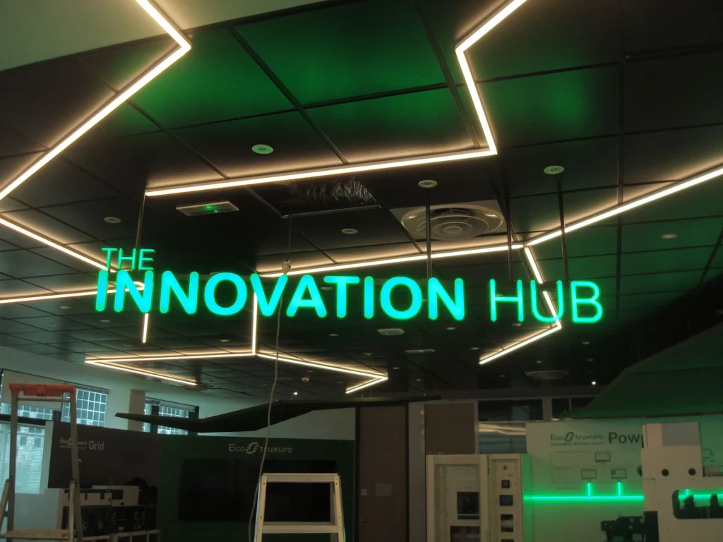 Enseigne lumineuse intérieure en lettres néon vert "Innovation Hub" suspendue sous un plafond technique design.